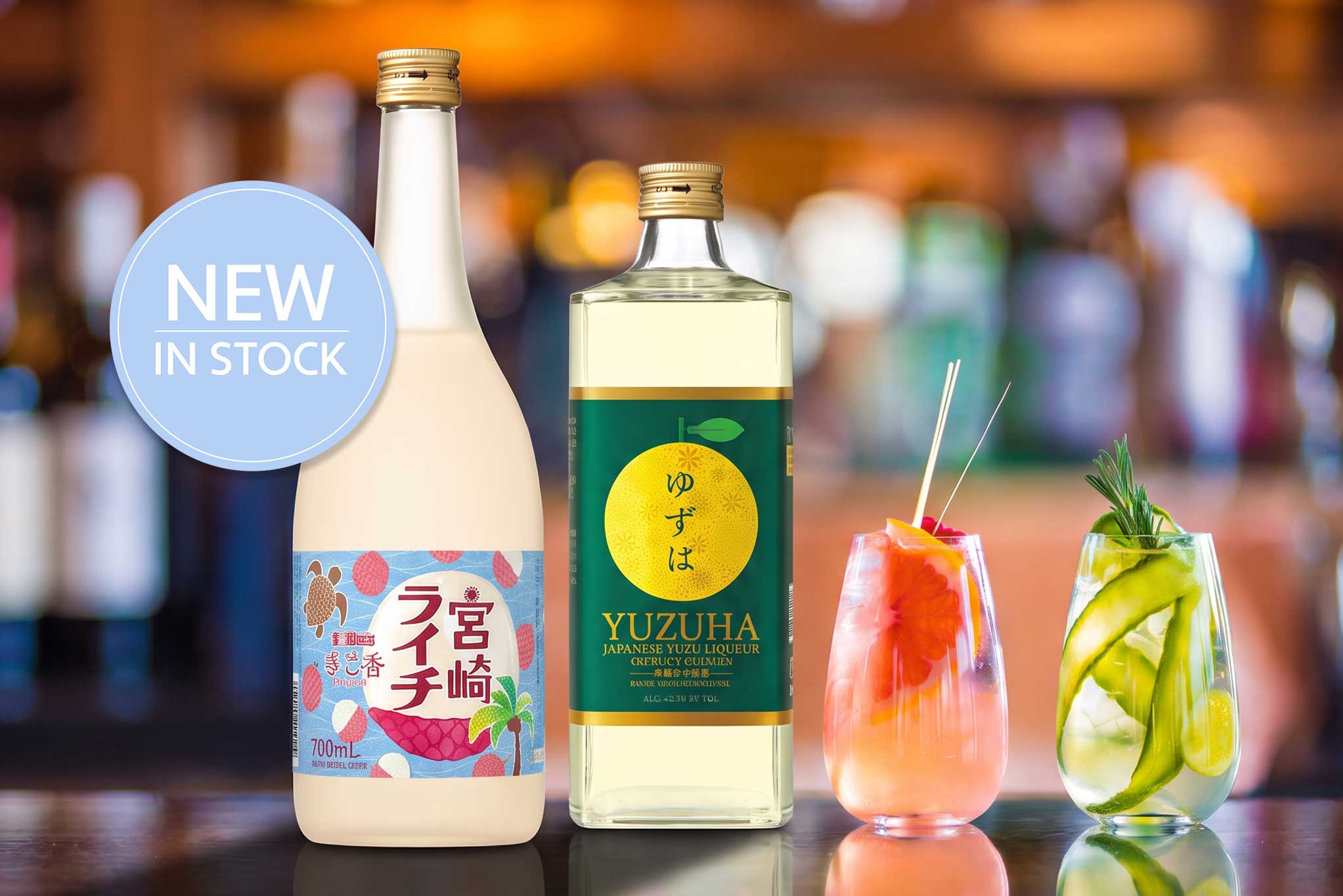 Zwei japanische Liköre auf einer Bar: Yuzuha Yuzu-Likör und cremiger Ananas-Litschi-Likör, daneben zwei dekorierte Cocktails und Hinweis "New in Stock"